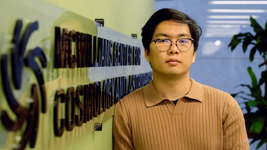 Man with glasses wearing beige collared shirt stands near a wall with a marquee that says "McWilliams Center for Cosmology and Astrophysics" in black letters