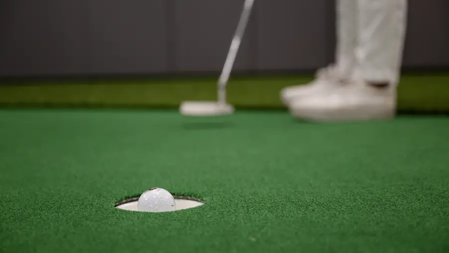 A close-up photo of a golf ball about to sink in to a putting hole shows the golfer's feet and club in the background after taking the shot.