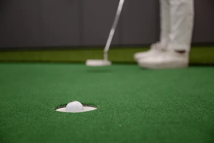A close-up photo of a golf ball about to sink in to a putting hole shows the golfer's feet and club in the background after taking the shot.